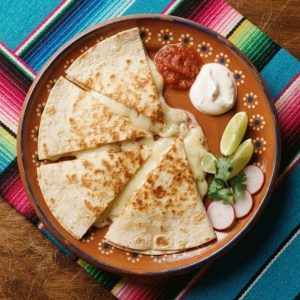 A flour tortilla quesadilla cut into wedges showing melted cheese, with salsa and sour cream, on a terracotta clay plate.